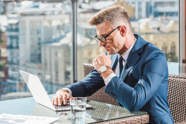 Vista lateral del hombre de negocios enfocado en anteojos usando computadora portátil en la mesa con vaso de agua en la cafetería - foto de stock