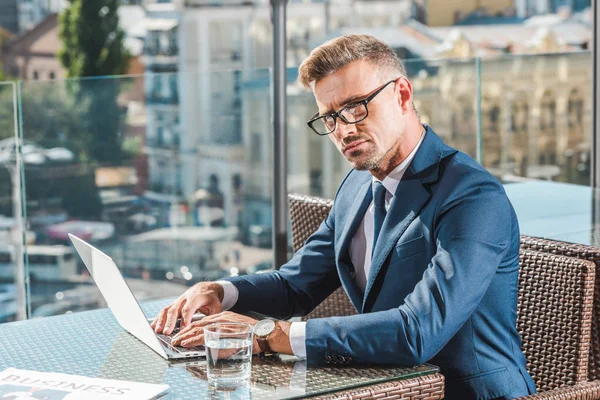 Confident businessman in eyeglasses sitting at table with laptop and glass of water in cafe — Stock Photo