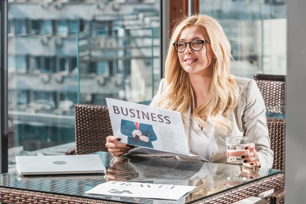 Retrato de mulher de negócios sorridente com vidro de jornal de leitura de água à mesa no café — Fotografia de Stock