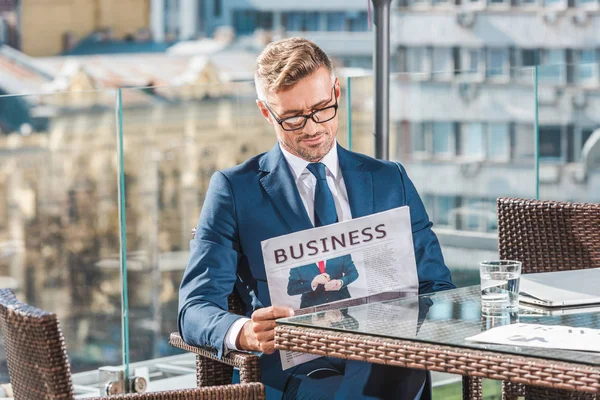 Portrait of stylish businessman in suit and eyeglasses reading newspaper in cafe — Stock Photo