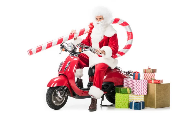 Серьезный Санта-Клаус в костюме, держащий большую рождественскую палку и сидящий на скутере рядом с кучей подарочных коробок, изолированных на белом фоне — стоковое фото