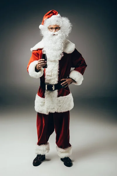 Happy santa claus in costume standing with cream soda bottle on grey background — Stock Photo