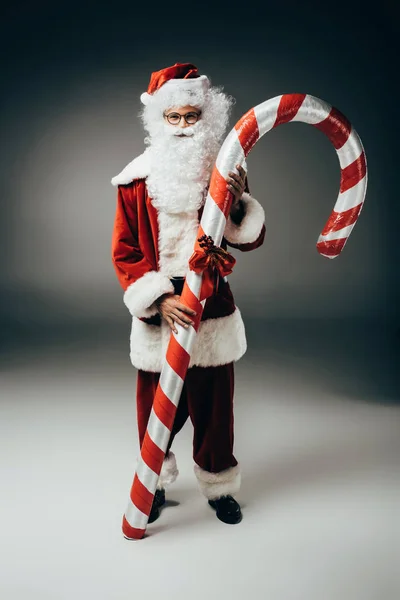 Feliz santa claus en traje de pie con gran palo de Navidad rayado sobre fondo gris - foto de stock