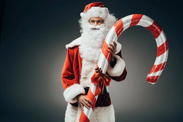Santa claus rêveur en costume debout avec gros bâton de Noël rayé isolé sur fond gris — Photo de stock