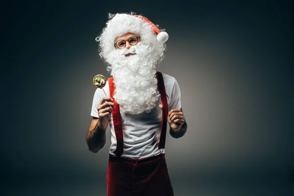 Fröhlicher Weihnachtsmann in Brille mit buntem Lutscher auf grauem Hintergrund — Stockfoto