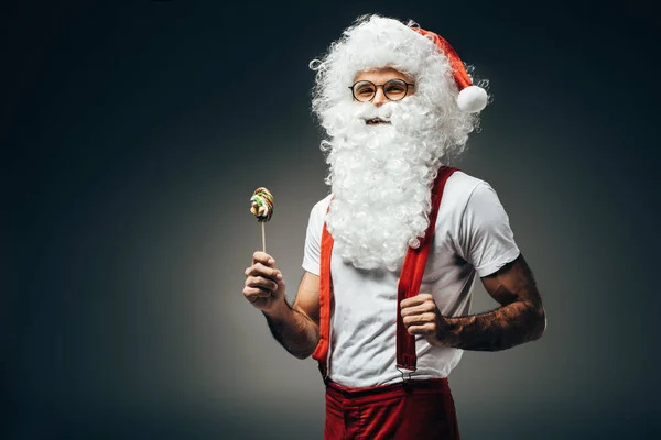 Lächelnder Weihnachtsmann in Brille mit buntem Lutscher auf grauem Hintergrund — Stockfoto