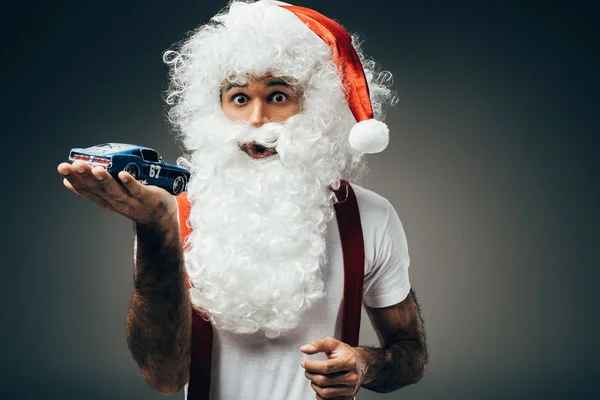 Schockierter Weihnachtsmann hält Spielzeugauto auf Handfläche und steht isoliert auf grauem Hintergrund — Stockfoto