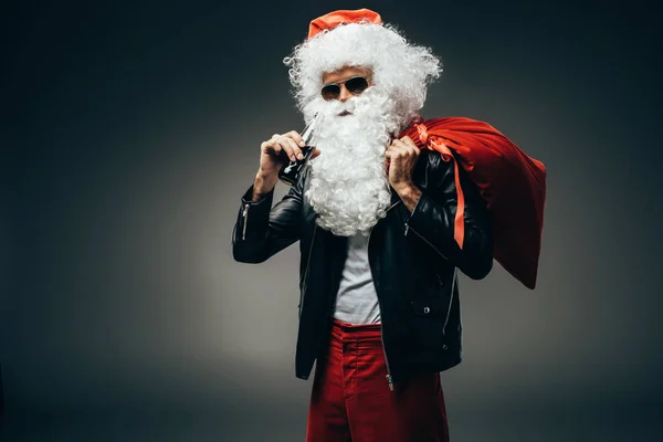 Stylish santa claus in sunglasses and leather jacket drinking cream soda and holding christmas bag over shoulder isolated on grey background — Stock Photo