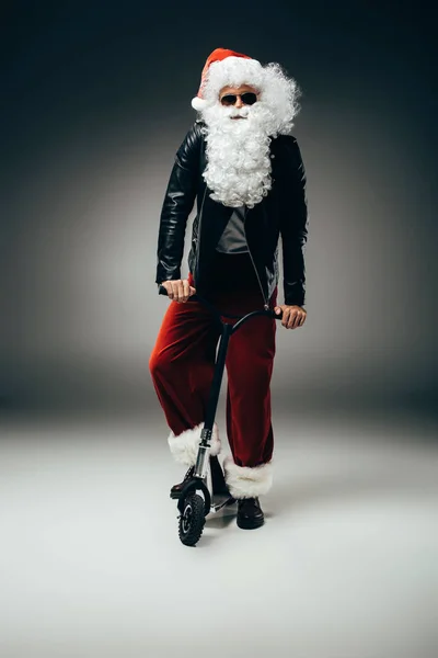 Sérieux santa claus en lunettes de soleil et veste en cuir debout avec trottinette de pied sur fond gris — Photo de stock