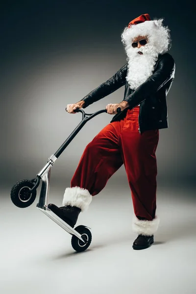 Drôle santa claus en lunettes de soleil et veste en cuir debout avec scooter kick sur fond gris — Photo de stock