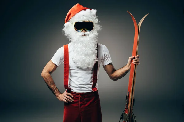 Santa claus sérieux dans le masque de ski debout avec la main sur la taille et tenant des skis isolés sur fond gris — Photo de stock