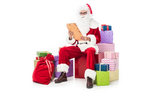 Papai Noel em óculos lendo pergaminho velho e sentado em pilha de caixas de presente isolado em fundo branco — Fotografia de Stock