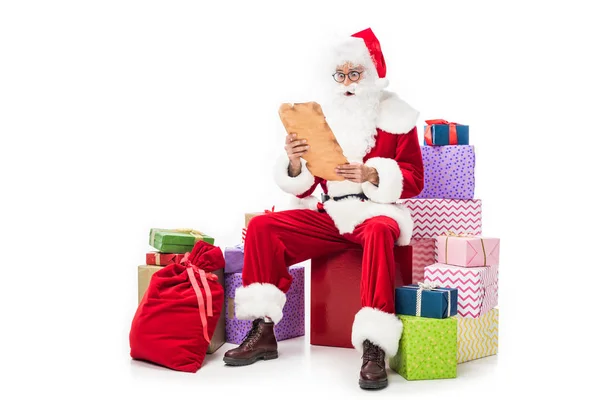 Impactado Santa Claus en anteojos leyendo pergamino viejo y sentado en un montón de cajas de regalo aislados sobre fondo blanco - foto de stock