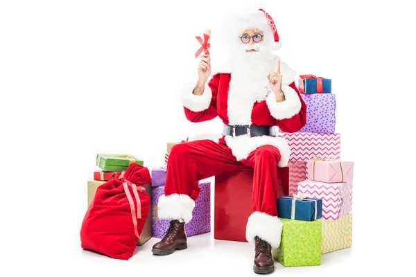 Santa claus faire geste idée et tenant cadeau de Noël tout en étant assis sur une pile de boîtes-cadeaux isolé sur fond blanc — Photo de stock