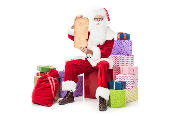 Санта-Клаус показує порожній старий пергамент і сидить на купі подарункових коробок ізольовані на білому тлі — стокове фото