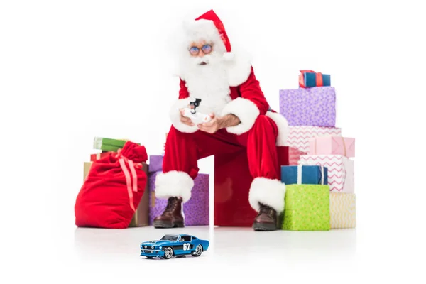 Santa claus sentado em pilha de caixas de presente e usando rádio controlado carro isolado no fundo branco — Fotografia de Stock