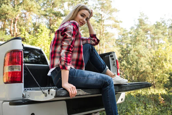 Low angle view of smiling woman looking at camera and sitting in trunk of pick up car outdoors — Stock Photo