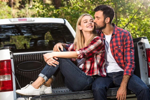 Handsome young man kissing attractive girlfriend while sitting on car trunk outdoors — Stock Photo