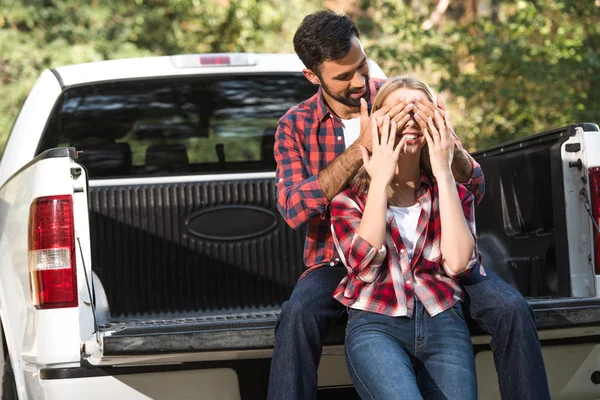 Jovem homem cobrindo olhos de namorada por trás enquanto sentado no porta-malas do carro ao ar livre — Fotografia de Stock