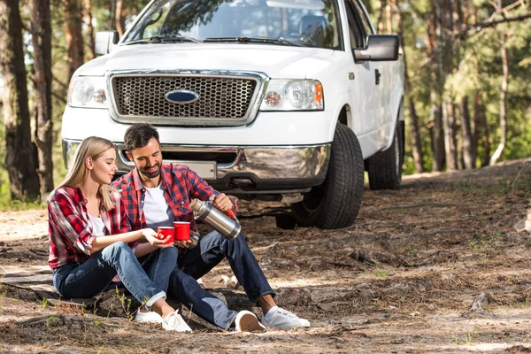 Sorrindo homem derramando café para namorada xícara de garrafa térmica perto pegar carro ao ar livre — Fotografia de Stock
