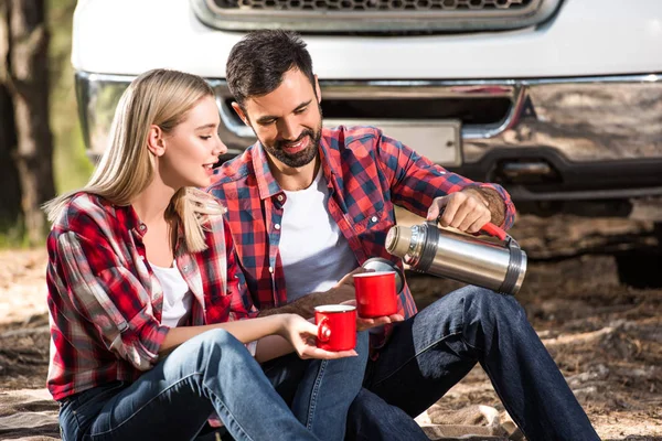Jovem feliz derramando café para o copo namorada de garrafa térmica perto do carro ao ar livre — Fotografia de Stock