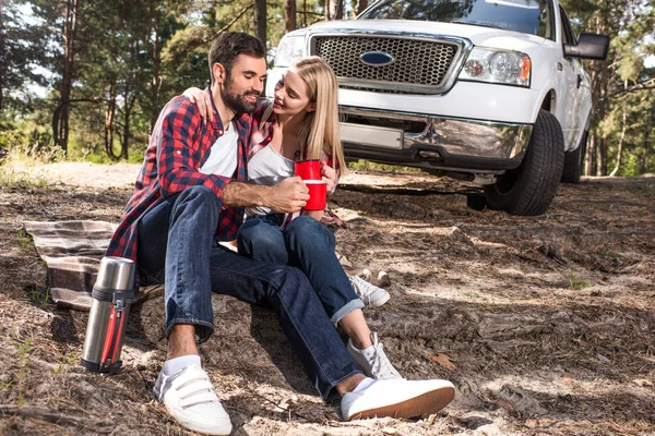 Casal alegre sentado no chão e clinking por copos de café perto pegar carro na floresta — Fotografia de Stock