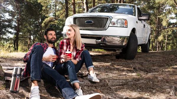 Sorrindo jovem casal sentado no chão com xícaras de café perto pegar carro na floresta — Fotografia de Stock