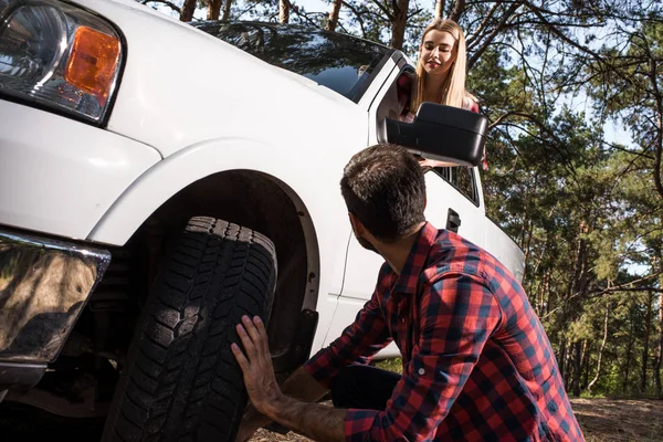 Rear view of man fixing wheel while his girlfriend sitting inside of pick up car outdoors — Stock Photo