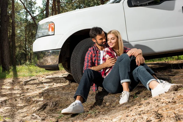 Молодая пара сидит на земле и обнимается рядом подобрать автомобиль на открытом воздухе — стоковое фото