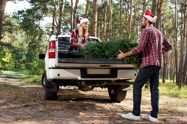 Jovem casal em santa chapéus carregando árvore de natal no carro tronco na floresta — Fotografia de Stock