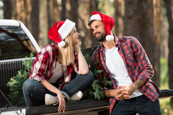 Улыбающаяся пара в рождественских шляпах, смотрящая друг на друга возле елки в багажнике машины — стоковое фото