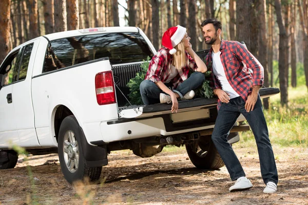 Beautiful woman in santa hat talking with boyfriend while sitting in car trunk with christmas tree in forest — Stock Photo