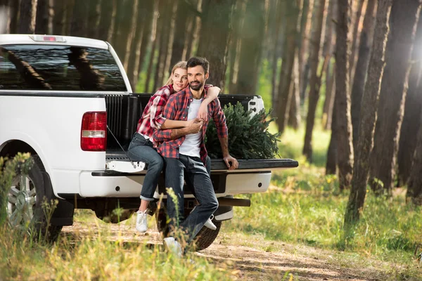 Bella giovane donna abbracciare fidanzato da dietro mentre seduto in auto tronco con albero di Natale nella foresta — Foto stock