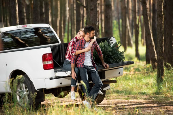 Enfoque selectivo de la hermosa mujer joven abrazando novio por detrás mientras está sentado en el tronco del coche con árbol de Navidad en el bosque - foto de stock
