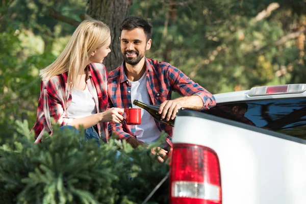 Homme heureux versant champagne à petite amie dans la tasse tout en étant assis dans le coffre de la voiture avec arbre de Noël dans la forêt — Photo de stock