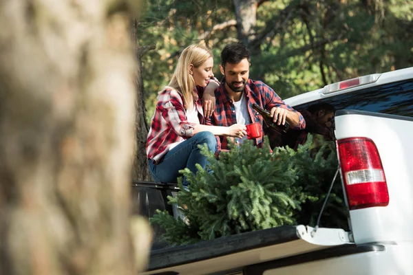 Selective focus of man pouring champagne to girlfriend into cup while sitting in car trunk with christmas tree in forest — Stock Photo