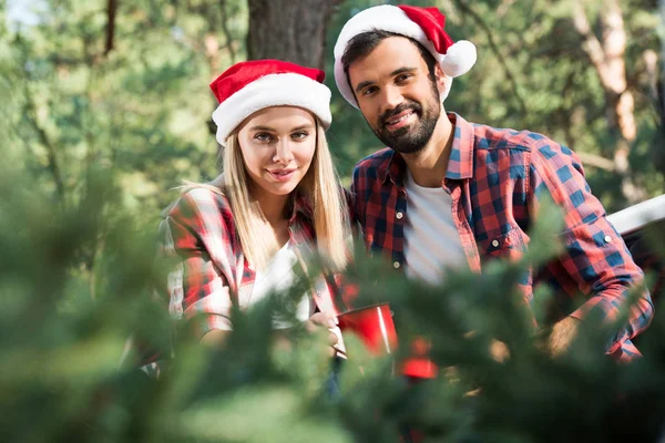 Selektiver Fokus des jungen Paares in Weihnachtsmützen mit Tassen und Blick in die Kamera — Stockfoto