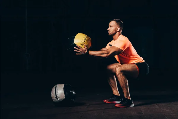 Handsome athletic sportsman doing squats with medicine ball in dark gym — Stock Photo