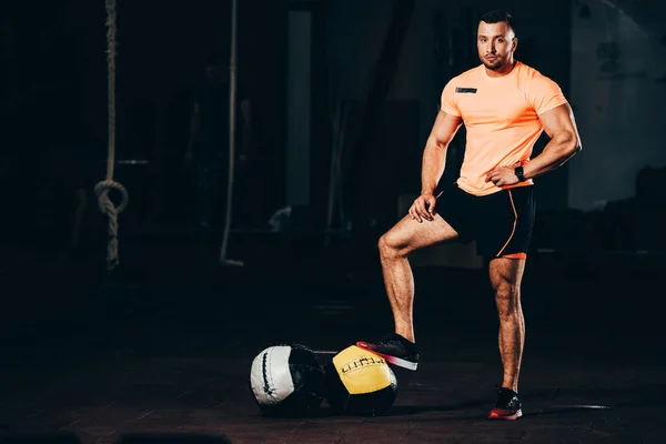 Schöner athletischer Bodybuilder steht in dunkler Turnhalle mit Medizinbällen am Boden und blickt in die Kamera — Stockfoto