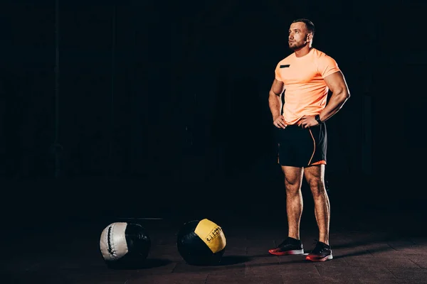 Apuesto atlético culturista de pie en el gimnasio oscuro con bolas de medicina en el suelo - foto de stock