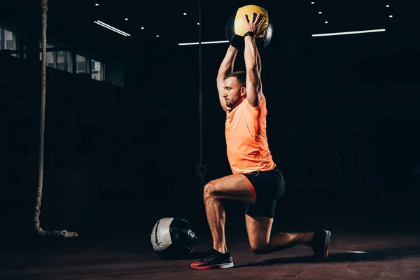 Handsome fit sportsman performing lunge with medicine ball overhead in dark gym — Stock Photo