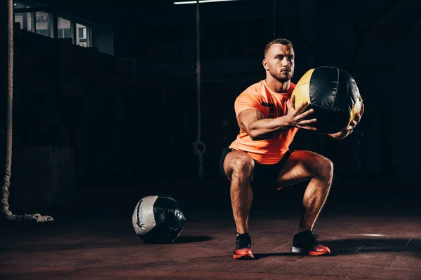 Beau sportif sportif effectuant squats avec boule de médecine dans la salle de gym sombre — Photo de stock