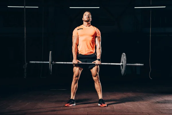 Hombre atlético guapo haciendo ejercicio con barra en el gimnasio oscuro y mirando hacia arriba - foto de stock