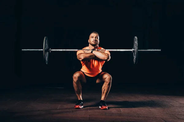 Handsome muscular man doing squats with barbell in dark gym — Stock Photo