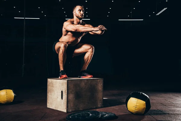 Beau bodybuilder athlétique faisant squats sur cube dans la salle de gym sombre — Photo de stock