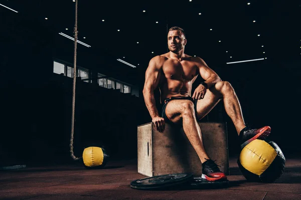 Deportista atlético sin camisa sentado en el cubo en el gimnasio oscuro - foto de stock