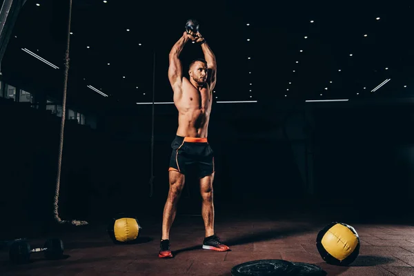 Apuesto deportista en forma sosteniendo kettlebell sobre la cabeza mientras se entrena en el gimnasio oscuro - foto de stock