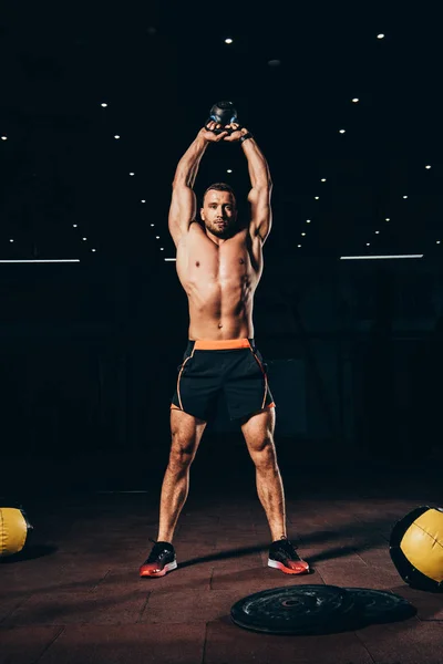 Handsome muscular man holding kettlebell overhead while working out  in dark gym — Stock Photo