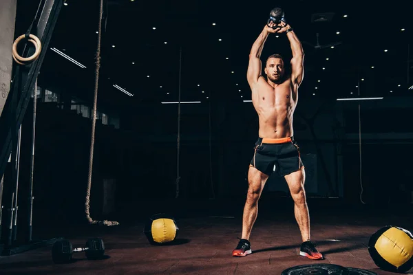 Handsome athletic man holding kettlebell overhead while working out  in dark gym — Stock Photo