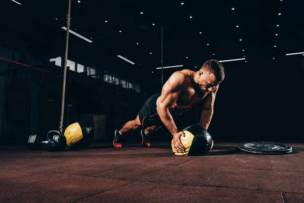 Apuesto deportista en forma haciendo flexiones en la pelota de medicina en el gimnasio oscuro - foto de stock